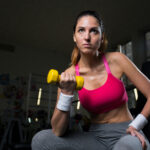 Young female in activewear sitting in gym and exercising with dumbbells